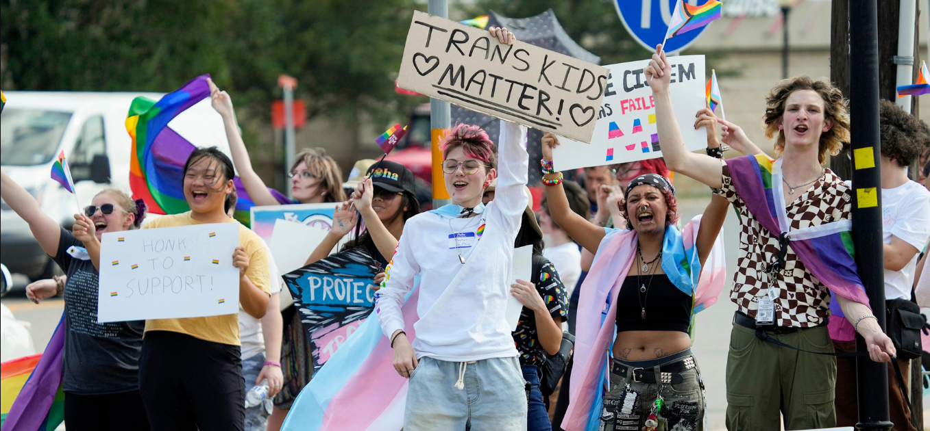 Pride march participants holding signs reading 'Trans Kids Matter' and 'Honk to Support'